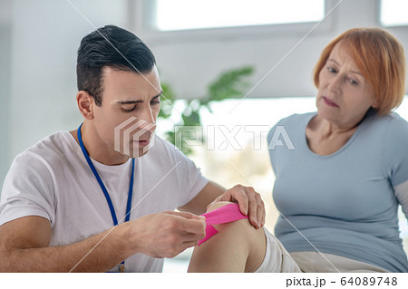 Skilled male therapist putting a special band on the knee 64089748