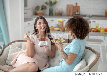 Smiling woman eating chocolate, sad girlfriend with plate of salad. 64090749