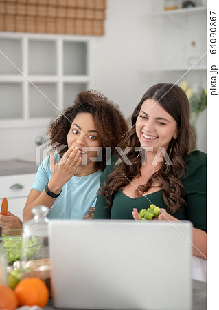 Two young women girlfriends in kitchen in front of laptop. Two young women girlfriends in kitchen in front of laptop. 64090867