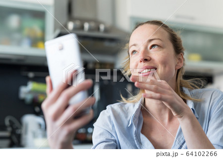 Young smiling cheerful pleased woman indoors at home kitchen using social media apps on mobile phone for chatting and stying connected with her loved ones. Stay at home, social distancing lifestyle. 64102266