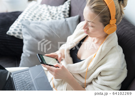 Social distancing. Stay at home. Woman in bathrobe being comfortable at her home sofa, using social media apps on phone for video chatting and stying connected with her loved ones. 64102269