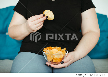Obese woman sitting on sofa eating unhealthy food 64109757