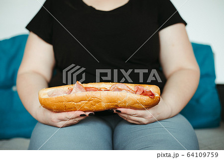 Obese woman sitting on sofa eating unhealthy food 64109759