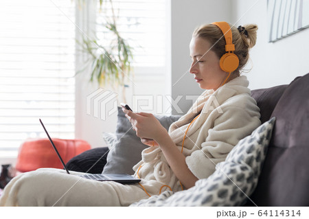 Social distancing. Stay at home. Woman in bathrobe being comfortable at her home sofa, using social media apps on phone for video chatting and stying connected with her loved ones. Social distancing. Stay at home. Woman in bathrobe being comfortable at her home sofa, using social media apps on phone for video chatting and stying connected with her loved ones. 64114314