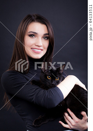 young brunette woman in black clothes on a black background with a black cat 64133511