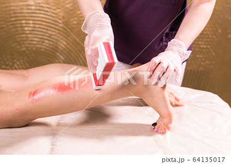 Waxing woman leg. Salon wax beautician epilation procedure. Waxing female body for hair removal by therapist close up. Smooth legs concept. Waxing woman leg. Salon wax beautician epilation procedure. Waxing female body for hair removal by therapist close up. Smooth legs concept. 64135017