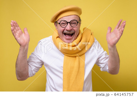 Surprised senior man in yellow scarf and beret holding hands up and smiling Surprised senior man in yellow scarf and beret holding hands up and smiling 64145757