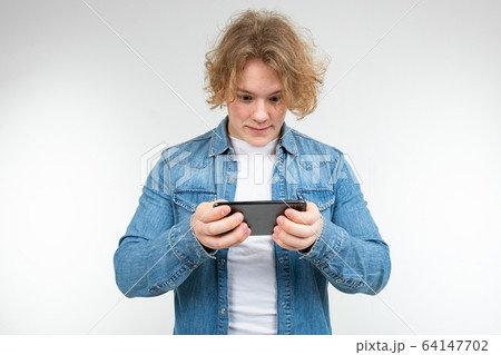 male gamer with blond hair in a denim jacket carefully plays on a smartphone on a white background 64147702