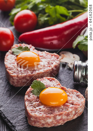 raw heart burger with egg on a black background with vegetables and herbs. 64162068