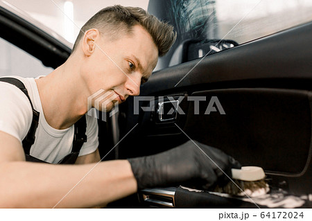 Close up portrait of professional car wash service worker in black protective gloves, cleaning car interior with special chemical foam and soft brush. Selective focus on worker face Close up portrait of professional car wash service worker in black protective gloves, cleaning car interior with special chemical foam and soft brush. Selective focus on worker face 64172024