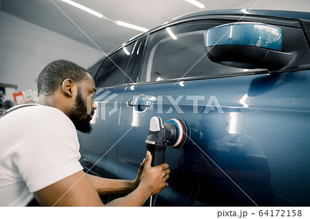 Young African man working at car detailing service, polishing blue car with polisher to eliminate contaminants from the surface of the car 64172158