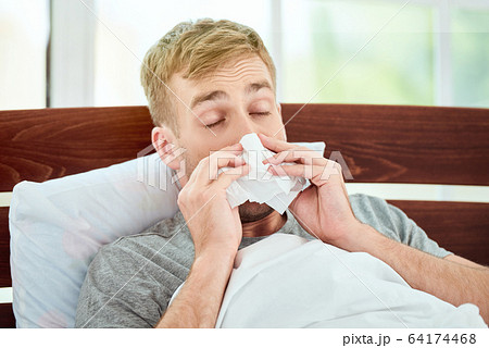 Portrait of a sick young man with runny nose suffering from cold or flu and sneezing while lying in bed at home. Virus disease 64174468
