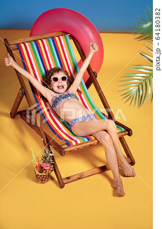 Smiling girl in swimsuit and sunglasses lying in rainbow deck chair with hands raised up emotionally and sunbathing 64180382