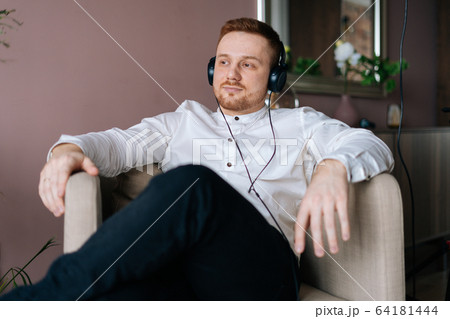 Bearded young man in headphones listening music, sitting on soft white armchair in living room. Bearded young man in headphones listening music, sitting on soft white armchair in living room. 64181444