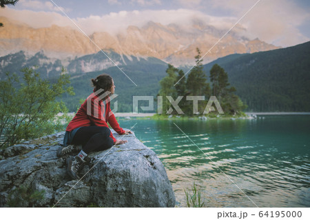 Germany, Bavaria, Eibsee, Young woman sitting on rock and looking at scenic view by Eibsee lake in Bavarian Alps 64195000