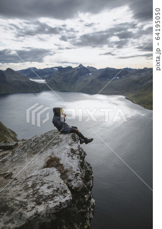 Norway, Senja, Man taking photo sitting on edge of steep cliff on top of mountain Segla Norway, Senja, Man taking photo sitting on edge of steep cliff on top of mountain Segla 64195050