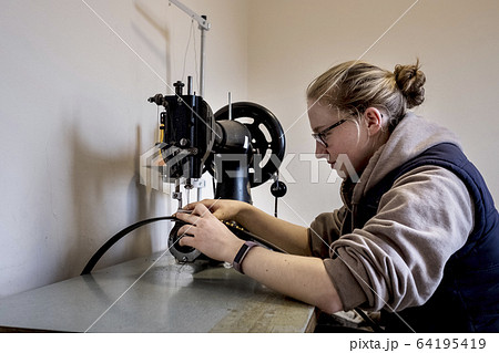 Female saddler sitting in workshop, sewing on saddlery sewing machine. Female saddler sitting in workshop, sewing on saddlery sewing machine. 64195419