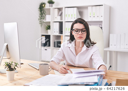 Young serious businesswoman looking at financial paper while making notes 64217626