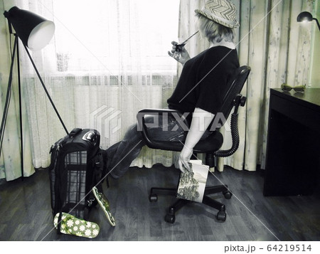 awaiting vacation during quarantine. Quarantine. Self isolation. Stay at home. A woman in a chair with a cocktail and a book looks out the window. Nearby a suitcase packed for vacation. 64219514