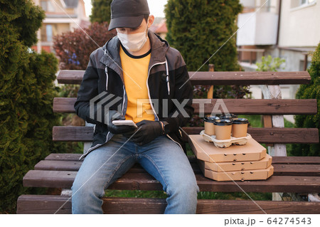 Courier in protective mask and medical gloves delivers pizza and coffe. Delivery boy sit on bench call to customer. Delivery service under quarantine. Coronavirus covid-19 theme Courier in protective mask and medical gloves delivers pizza and coffe. Delivery boy sit on bench call to customer. Delivery service under quarantine. Coronavirus covid-19 theme 64274543