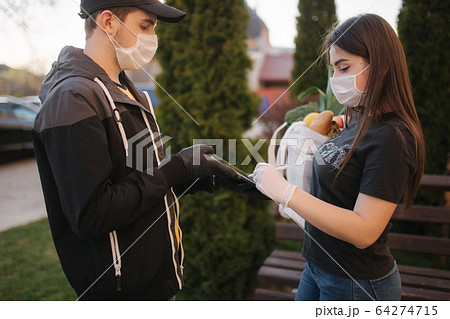 Smart food delivery service. Man in medical mask and gloves handing fresh food to young woman customer receiving express delivery from courier at home. Courier with tablet, sign in. Female order food 64274715