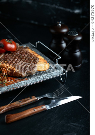 Grilled steak with knife and fork carved on black stone slate. Steak on a hot marble stone. Copy space, dark background, food fashion photo. Grilled steak with knife and fork carved on black stone slate. Steak on a hot marble stone. Copy space, dark background, food fashion photo. 64275970