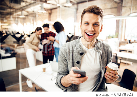 Cheerful young man holding smartphone and cup of coffee Cheerful young man holding smartphone and cup of coffee 64279923