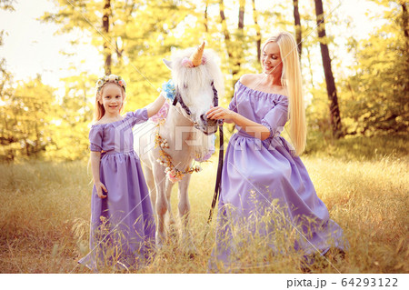 Mother and daughter in similar lavender dresses are petting a unicorn horse. Summer meadow 64293122