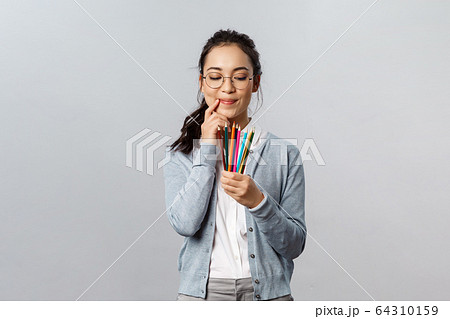 Hobbies, creativity and art concept. Thoughtful young creative female artist touch lip thinking and smiling as look at colored pencils, plan what to draw next for inktober, stand grey background 64310159
