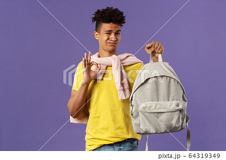 Good but not the best. Portrait of disappointed, unsatisfied young teenage student, guy grimacing without enthusiasm show okay, received new ugly backpack, standing purple background 64319349
