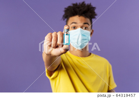 Medicine, coronavirus and people concept. Close-up portrait of young man in facial mask holding ampoule with drug, finally found vaccine from covid19, standing purple background Medicine, coronavirus and people concept. Close-up portrait of young man in facial mask holding ampoule with drug, finally found vaccine from covid19, standing purple background 64319457