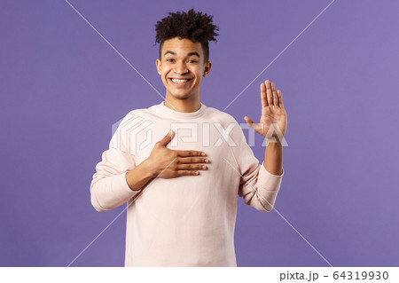 Portrait of young happy, joyful hispanic man making honest statement, promise to tell only truth, hold one hand on heart and another raised, smiling sincere, standing purple background 64319930