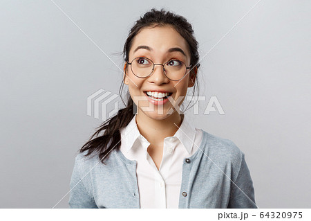 Close-up portrait of upbeat, excited and amused asian woman in glasses, look intrigued at upper left corner, reading interesting proposal or chart, standing grey background astonished 64320975