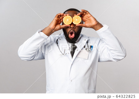 Healthcare, healthy diet and disease concept. Funny good-looking physician, african-american doctor playing with orange, look amused, tell stay safe, eat vitamins and fruits, grey background 64322825