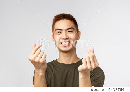 People, different expressions and lifestyle concept. Cute asian guy with stylish haircut, showing korean finger hearts and smiling lovely, express sympathy or like, stand grey background People, different expressions and lifestyle concept. Cute asian guy with stylish haircut, showing korean finger hearts and smiling lovely, express sympathy or like, stand grey background 64327644