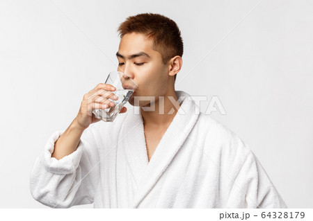 Beauty, spa and technology concept. Portrait of young handsome healthy asian man, drinking water from glass and wear bathrobe, standing white background, start morning with sip of aqua Beauty, spa and technology concept. Portrait of young handsome healthy asian man, drinking water from glass and wear bathrobe, standing white background, start morning with sip of aqua 64328179