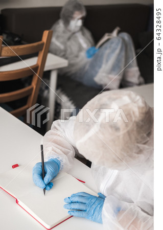 Girl in protective white translucent suit, blue rubber gloves, medical mask sits at table at home and draws in sketchbook with pencil. Remote work of designer, artist during coronavirus pandemic. Girl in protective white translucent suit, blue rubber gloves, medical mask sits at table at home and draws in sketchbook with pencil. Remote work of designer, artist during coronavirus pandemic. 64328495