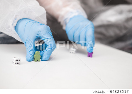 Two people in protective white translucent suit, blue rubber gloves, medical mask sit on sofa at home and play board games, roll dice. Stay home during coronavirus pandemic covid. 64328517
