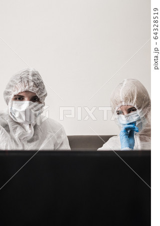 Two person in protective white translucent suit, blue rubber gloves, medical mask are sitting on sofa at home and watching the news on laptop. Stay home during coronavirus pandemic. 64328519
