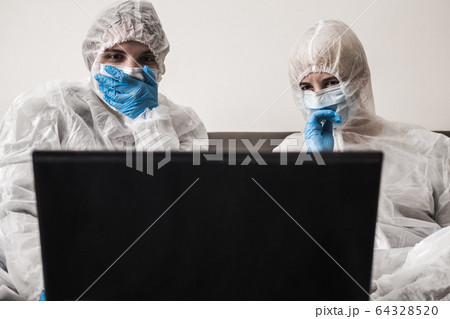 Two person in protective white translucent suit, blue rubber gloves, medical mask are sitting on sofa at home and watching the news on laptop. Stay home during coronavirus pandemic. Two person in protective white translucent suit, blue rubber gloves, medical mask are sitting on sofa at home and watching the news on laptop. Stay home during coronavirus pandemic. 64328520
