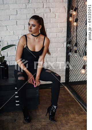 Beautiful fashionable woman at loft style room with white bricks wall and black iron net. Attractive girl with perfect figure in stylish black clothes. 64330341
