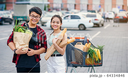 Likable asian couple hold paper eco bags with organic healthy food in hands while standing near store mall. Happy family shopping on weekend. Likable asian couple hold paper eco bags with organic healthy food in hands while standing near store mall. Happy family shopping on weekend. 64334846