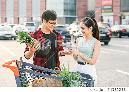 Smiling asian couple standing near shopping cart,man holds pineapple and cheerful woman counts the banknotes 64335258