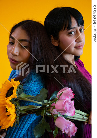 Two latin young ladies posing back to back, yellow background, roses and sunflowers 64344535