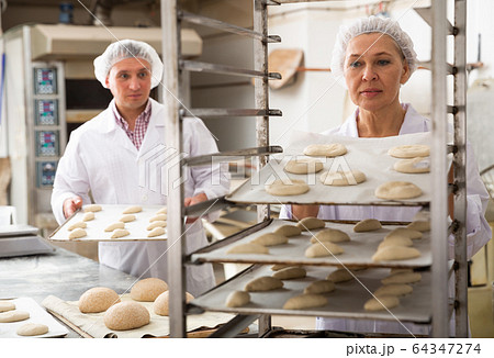 Female baker placing tray with formed dough on trolley 64347274