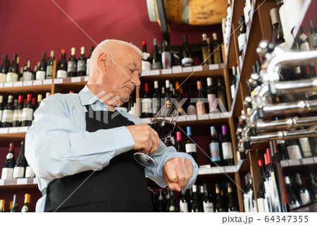 Confident elderly male winemaker holding glass of red wine, checking it in wine store 64347355
