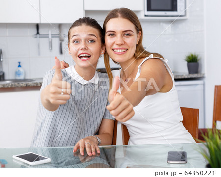 Young girlfriends sitting and holding thumbs up at kitchen table Young girlfriends sitting and holding thumbs up at kitchen table 64350221