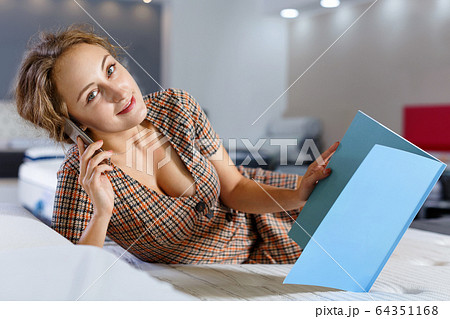 Young attractive woman lying on bed and talking on phone while choosing mattress Young attractive woman lying on bed and talking on phone while choosing mattress 64351168