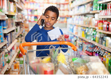 Focused African tween boy talking on phone and looking at shopping list while visiting supermarket Focused African tween boy talking on phone and looking at shopping list while visiting supermarket 64352243