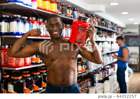 Bare-chested athletic African standing with cans of sports nutrition in store, demonstrating muscles 64352257